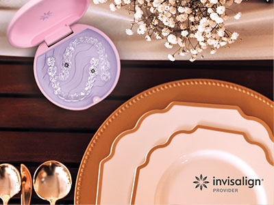 The image shows a table setting with various dinnerware items, including plates, bowls, and utensils, arranged on a wooden surface. There is also a decorative box with a floral design in the center of the table. In the background, there s a pink object that resembles a toothbrush holder with a toothbrush inside. The setting appears to be indoors, and there are some flowers visible behind the table. The image has text at the bottom right corner that reads  Invisalign    and  www.invisalign.com,  indicating the brand associated with the image.