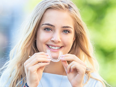 A young woman with blonde hair is smiling at the camera while holding a clear plastic dental retainer in her hand.