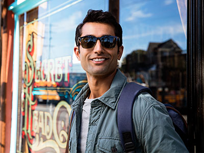 The image shows a man standing outdoors, wearing sunglasses and a backpack, posing in front of a store window with a sign that reads Salt Pepper .