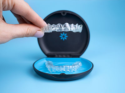 The image shows a person s hand holding an open box containing a set of clear plastic aligners, which are used for teeth straightening.