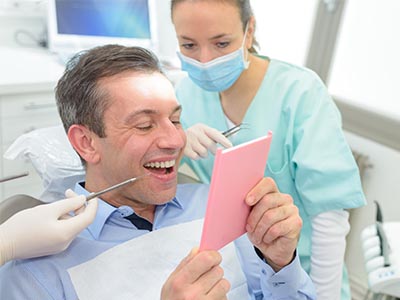 A man in a dental chair is holding up a pink card with a surprised expression, while a dentist and hygienist look on.
